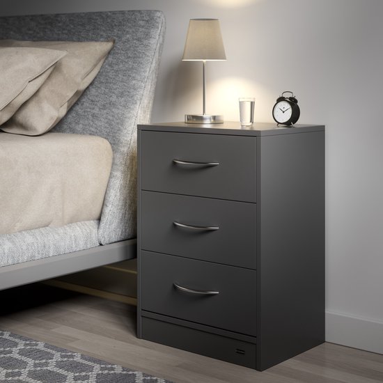Grijs nachtkastje met 3 lades, naast een bed met grijs gestoffeerd hoofdeinde en beige kussens. Een lampje, glas water en wekker staan erop.
