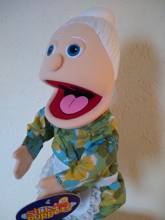 Close-up van een handpop van een oma met grote mond, blauwe ogen en wit haar. Draagt een groen met bloemenprint jurk en wit schort. Merk "Silly Puppets" zichtbaar.