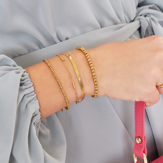 Arm van een vrouw met drie gouden armbanden: een met kleine kraaltjes, een platte schakelarmband met staafjes, en een met bolletjes.