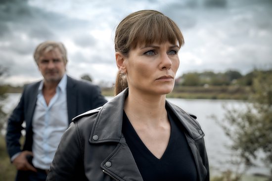 Een vrouw met bruin haar en een zwarte leren jas, met een man op de achtergrond.