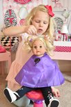 Een meisje met blonde haren, een roze strik en een lichtroze jurk speelt met een pop met blonde krullen, een paarse cape en kinderartikelen.