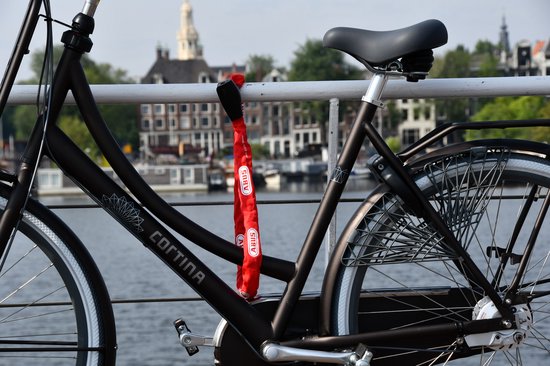 Fietsketting ABUS rood, bevestigd aan een fietsframe bij een waterkant met huizen op de achtergrond.