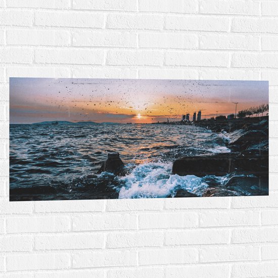 WallClassics - Muursticker - Klotsende Golven tegen Rotsen bij Zonsondergang - 100x50 cm Foto op Muursticker