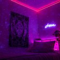 Een kamer met een bed, verlicht door een paarse sterrenhemel projectie en neonverlichting. Een wandkleed met zon en maan motief hangt aan de muur.