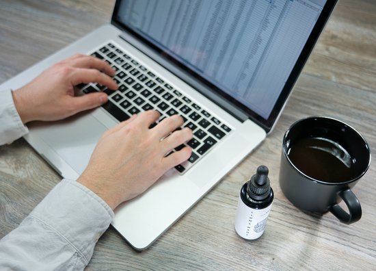 Iemand met witte blouse typt op een laptop, met een flesje tinctuur en zwarte mok ernaast.