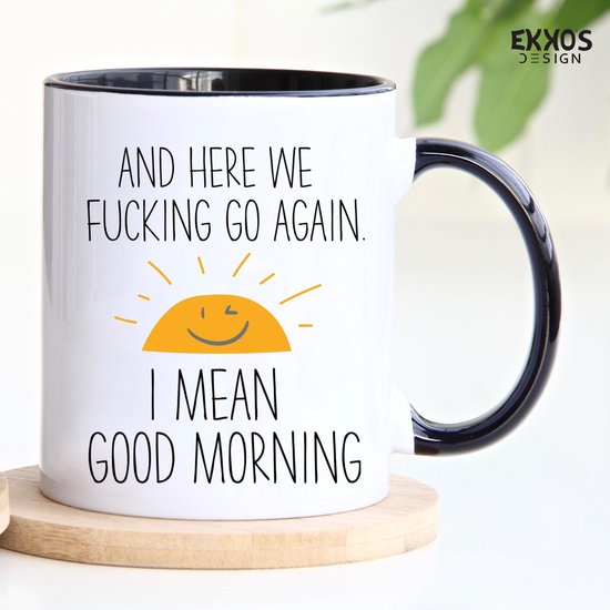 Witte mok met zwarte rand en oor, op een houten onderzetter. Tekst: "AND HERE WE FUCKING GO AGAIN. I MEAN GOOD MORNING" met lachende zon.