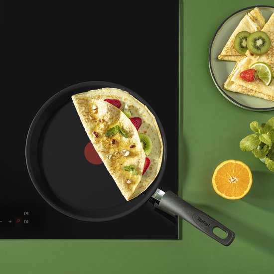 Zwarte Tefal crêpepan op een inductiekookplaat met een crêpe belegd met aardbeien, kiwi en noten.