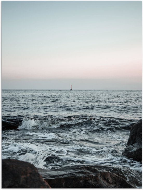 Foto van een woelige zee met rotsen op de voorgrond en een kleine rode vuurtoren aan de horizon.