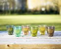 Six verres colorés sur une table en bois, remplis d'une boisson légère.