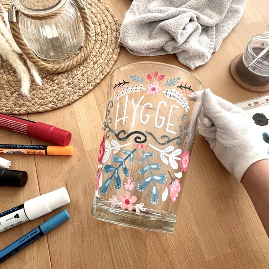 Un verre décoré de fleurs et du texte "HYGGE" en lettres blanches. Des décorations et des marqueurs sont posés sur la table.