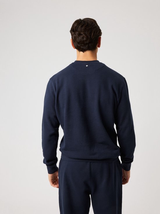 Achterkant van een donkerblauwe sweater met op de nek een klein wit logo. Model draagt de sweater.