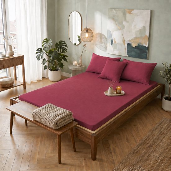Un lit fait avec du linge de lit bordeaux, des coussins et de la décoration dans une chambre lumineuse.