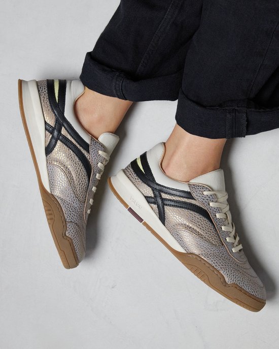 Luchtfoto van goudkleurige sneakers met reptielenprint, beige veters en zwarte accenten, gedragen door een persoon in een zwarte broek.