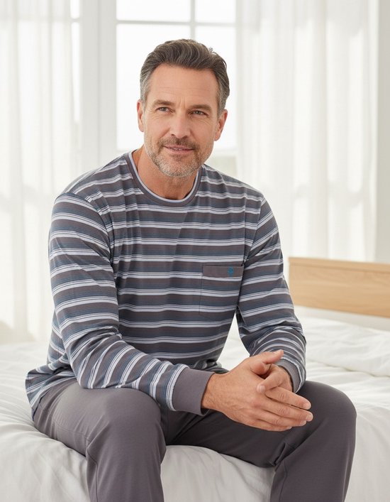 Man zittend op een bed in een gestreepte pyjamaset van Paul Hopkins. Grijze pyjamabroek met trekkoord.