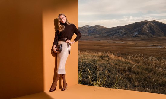 Model in een beige pailletten rok, bruine coltrui en bruine panty's, staand.