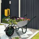 Groene kruiwagen gevuld met kleurrijke bloemen en zwarte grond, naast een schuur.