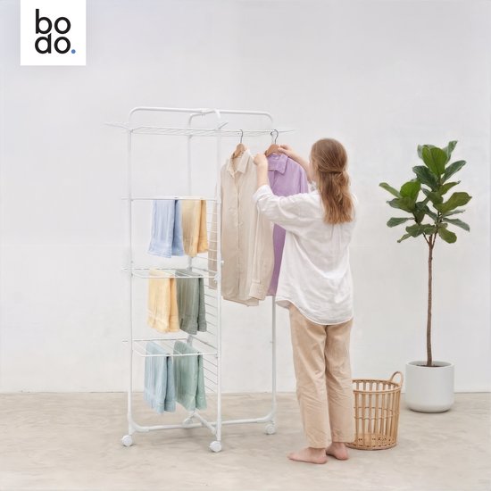 Een vrouw hangt kleding aan een witte droogtoren met vier etages in een lichte ruimte.