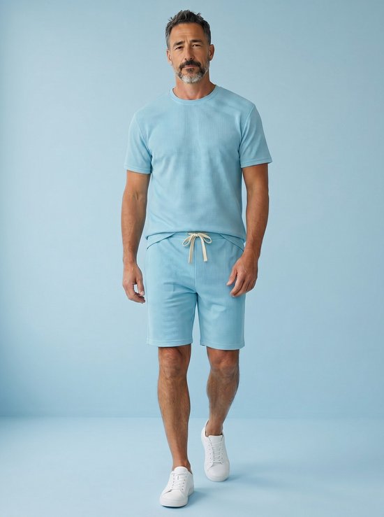Man met grijze baard loopt, draagt turquoise T-shirt en short met wafelpatroon en witte sneakers.
