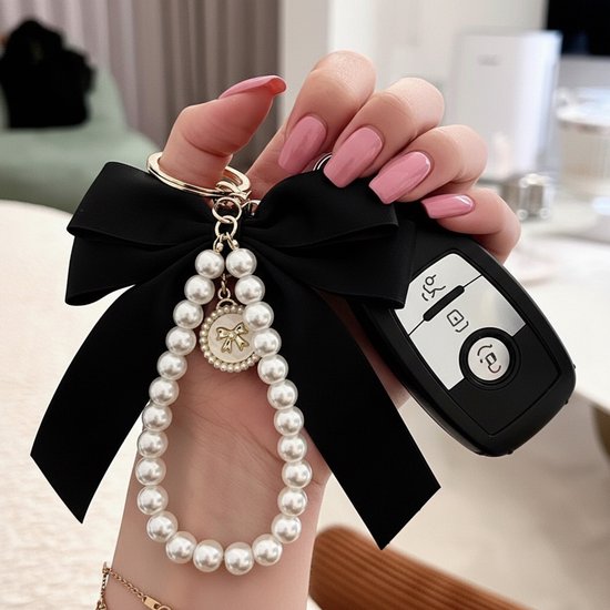 Hand met roze nagels houdt zwarte strik sleutelhanger met parelarmband en gouden strik-bedel vast, naast autosleutels.