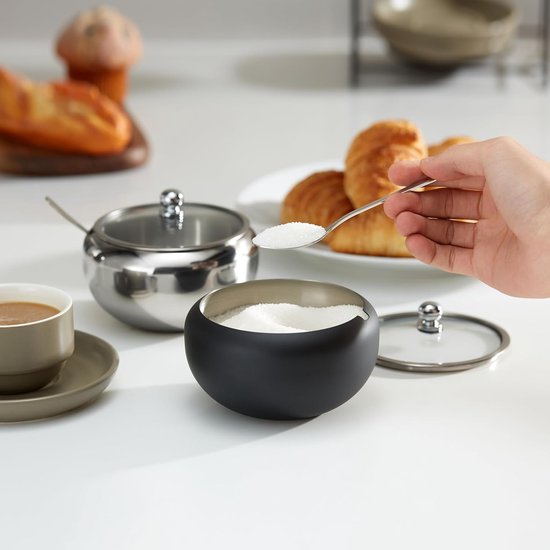 Zwarte suikerpot met glazen deksel, naast een RVS suikerpot en een kopje koffie.
