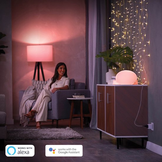 Een vrouw zit in een woonkamer, met "Works with Alexa" en "Works with Google Assistant" logo's, en een smart plug in de muur.