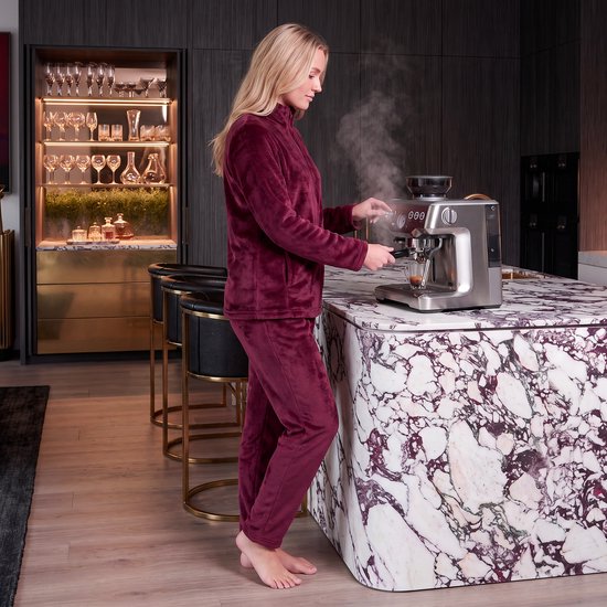 Een blonde vrouw in een bordeauxrood fleece huispak maakt koffie in een keuken.