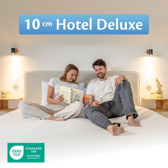 Deux personnes souriantes assises sur un lit avec un surmatelas Hotel Deluxe de 10 cm d'épaisseur. Homme avec une tasse, femme avec un livre. OEKO-TEX STANDARD 100.