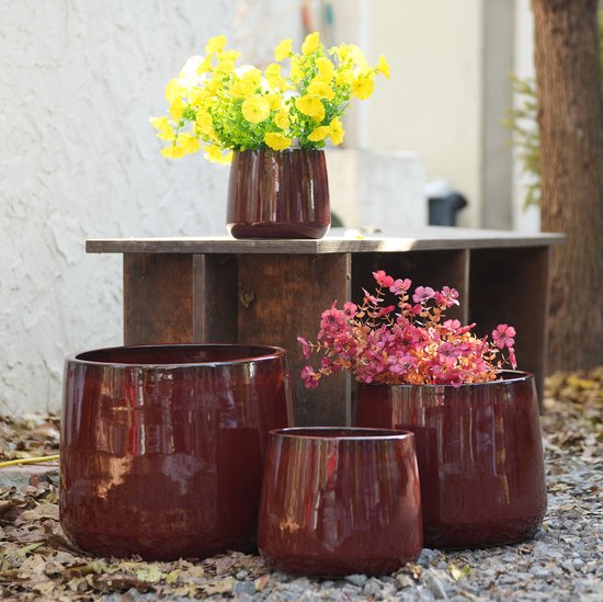 Set van 4 donkerrode, hoogglans keramieken bloempotten. Drie potten staan op de grond, de grootste met een brede opening. Eén pot staat op een houten tafel, gevuld met gele bloemen.
