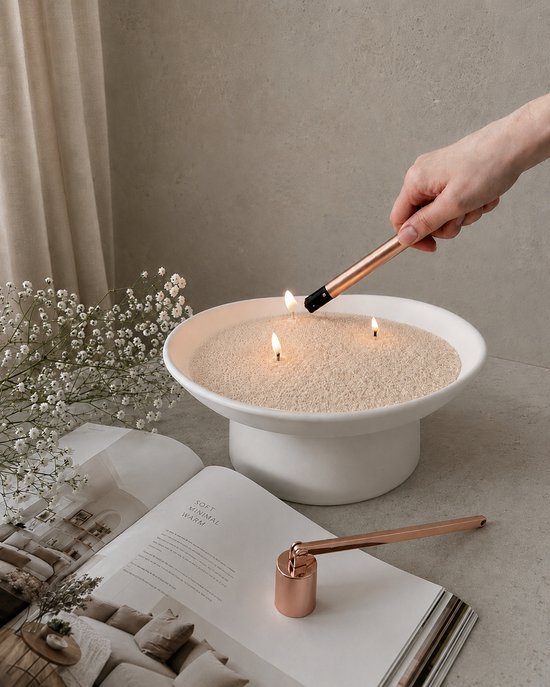 Brandende kaarsen in een witte schaal met beige kaarsenzand. Naast een open boek en een koperen lontknipper.