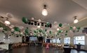 Een grote zaal versierd met witte en groene lampionnen. Houten tafels en stoelen staan verspreid.