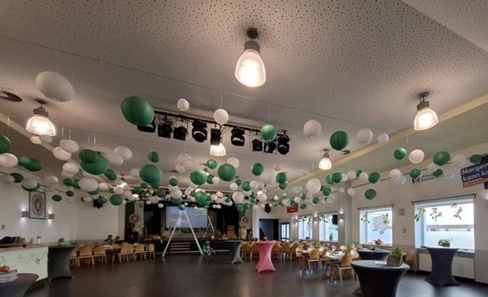 Een grote zaal versierd met witte en groene lampionnen. Houten tafels en stoelen staan verspreid.