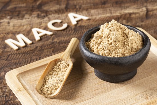 Maca poeder in een zwarte kom, met een houten schepje ernaast. Het woord 'MACA' is in houten letters op de achtergrond.