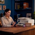 Vrouw zit aan een bureau met een HP printer en telefoon, met HP Instant Ink cartridges en een doosje.