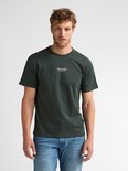 Donkergroen T-shirt met kleine witte tekstprint op de borst. Model draagt een spijkerbroek.