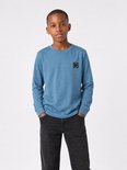 Jongen met handen in de zakken, draagt een blauw longsleeve T-shirt met borstlogo.