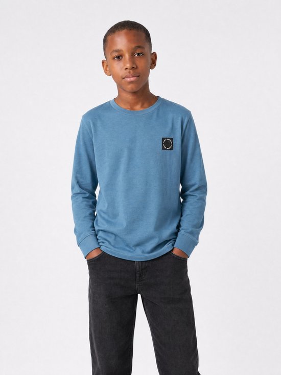 Jongen met handen in de zakken, draagt een blauw longsleeve T-shirt met borstlogo.