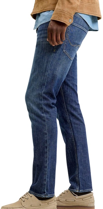 Man in een spijkerbroek, blauw overhemd en bruine jas, van de zijkant gezien.