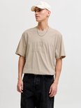 Man met beige T-shirt met "ORIGINALS" opdruk, zwart jeans en beige pet.