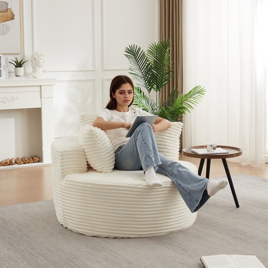 Een jonge vrouw zit op een crèmekleurige corduroy fauteuil met ribbelpatroon en drie kussens, met een tablet in haar handen.