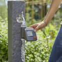 Een hand met jeans bedient een grijze GARDENA besproeiingscomputer met display, aangesloten op een kraan met stromend water.