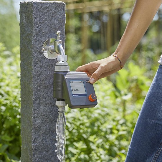 Een hand met jeans bedient een grijze GARDENA besproeiingscomputer met display, aangesloten op een kraan met stromend water.
