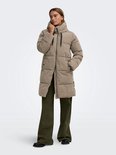Model in lange beige pufferjas met capuchon, zwarte koordjes en groene broek.