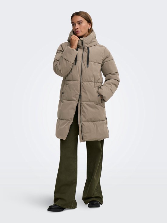 Model in lange beige pufferjas met capuchon, zwarte koordjes en groene broek.