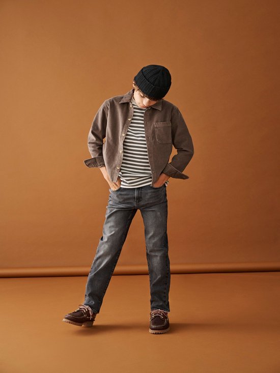 Jongen met zwart muts, gestreepte top, bruine jas en grijze jeans.