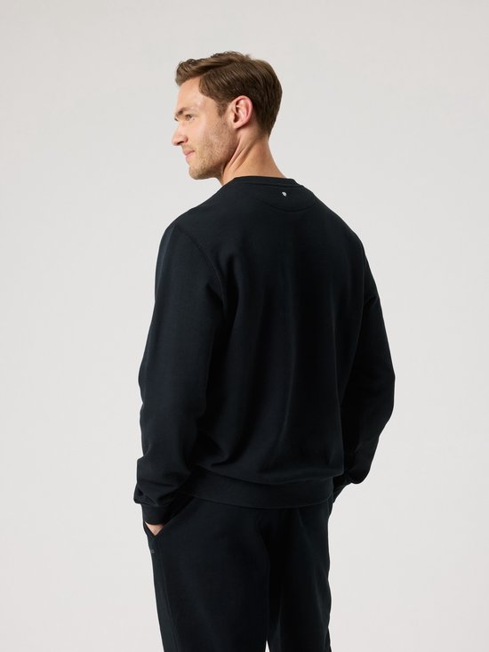 Man in een zwarte sweater, achterkant zichtbaar, met handen in zijn broekzakken.
