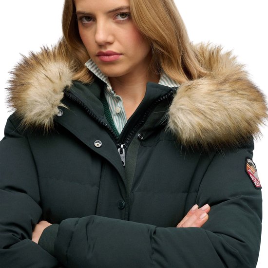 Gezicht van een vrouw met blond haar, draagt een donkergroene bomberjack met volle bontkraag en rits. Ze heeft een gestreepte blouse en kabeltrui aan, armen gekruist.