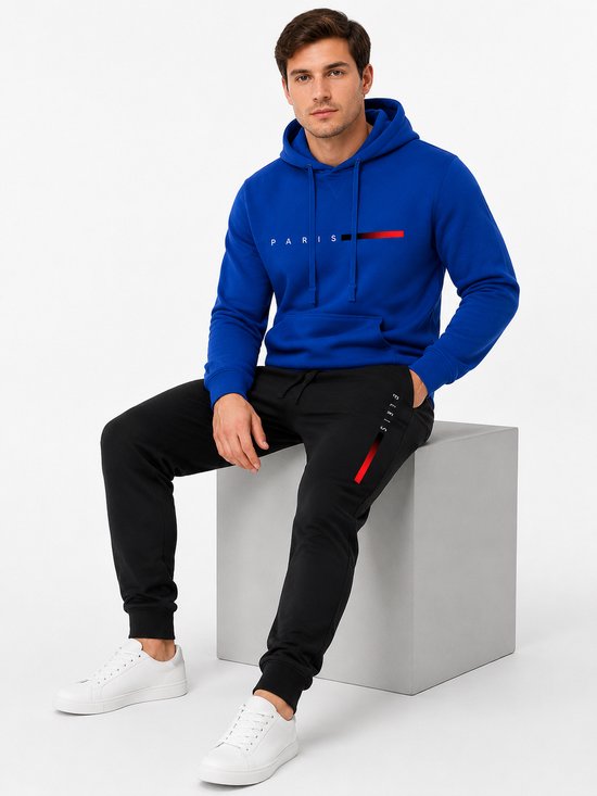 Man zit op een grijze kubus, draagt een blauwe hoodie met 'PARIS' tekst en rode/zwarte streepjes, en een zwarte joggingbroek.