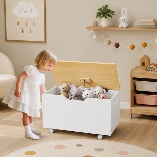 Wit speelgoedkist met houten deksel, gevuld met knuffels, met een jong kind dat ernaar kijkt.