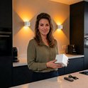 Femme dans une cuisine avec deux appliques murales blanches au mur projetant une lumière chaude. Elle tient une lampe carrée blanche.