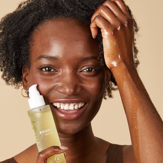 Vrolijke zwarte vrouw met krullend haar, houdt een fles 'BOUCLÈME Curls Redefined Revive 5 Hair Oil' vast.
